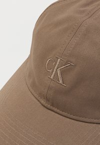 Căciulă maro din țesătură texturată, cu logo-ul brodat tonal "cK" pe față. Vizor curbat cu detalii vizibile de cusătură.