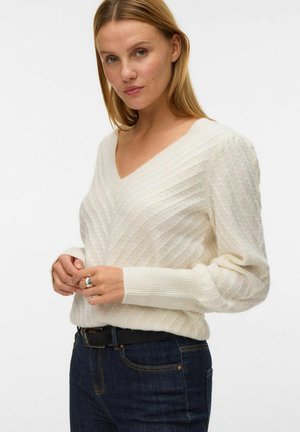 Sweter w kolorze kremowym z dekoltem w serek, z teksturowanym diagonalnym wzorem, ściągaczami na rękawach i luźnym krojem, połączony z ciemnoniebieskimi dżinsami i czarnym paskiem.