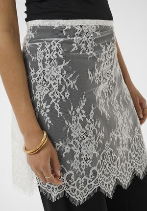 Jupe en dentelle florale blanche sur un pantalon noir portée par une personne avec des bracelets et des bagues en or à la main sur un fond uni.