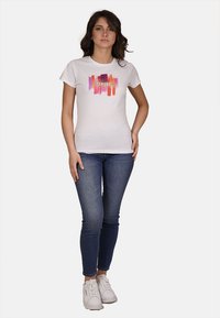 T-shirt bianco in cotone con un design astratto colorato e la scritta "YES-ZEE" al centro. Abbinato a jeans blu e sneakers bianche.