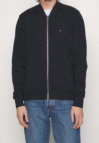 Persona con una chaqueta de zip azul marino con un pequeño logo en el pecho y jeans azules, posando frente a un fondo liso.