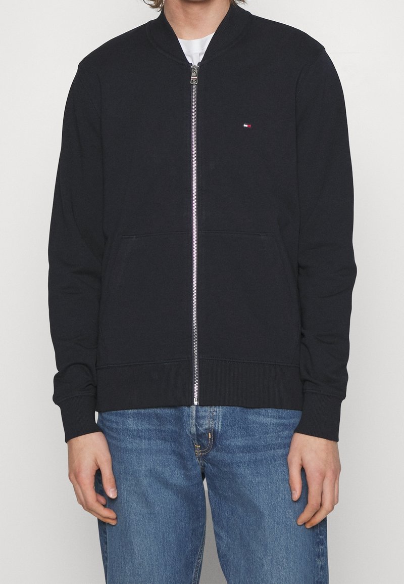 Persona con una chaqueta de zip azul marino con un pequeño logo en el pecho y jeans azules, posando frente a un fondo liso.