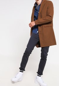 Cappotto lungo in misto lana marrone, jeans grigi, sneakers bianche con accenti neri, indossato sopra una camicia blu a bottoni, design e vestibilità casual.