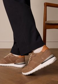 Bruine suède sneakers met witte zolen, uitgerust met een lichtbruine accent, beige veters en een gestructureerd oppervlak, gecombineerd met donkere broeken.