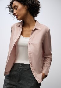 Chemise rose clair, à boutons, avec un col classique, des manches longues et une texture lisse, portée sur un débardeur blanc et un pantalon gris texturé.