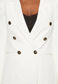 Blazer bianco doppiopetto con bottoni decorati in oro e nero, caratterizzato da revers a punta e tasche laterali, abbinato a un top in pizzo nero sotto.