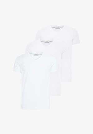 Jeune homme aux cheveux bouclés debout face à l'avant, portant un T-shirt blanc à col en V, un pantalon plissé gris et des baskets blanches avec des rayures noires.