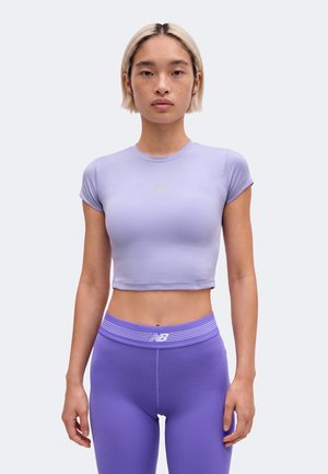 Ung kvinde iført en lyse lilla cropped T-shirt og matchende højtaljede leggings, stående mod en ensfarvet baggrund.
