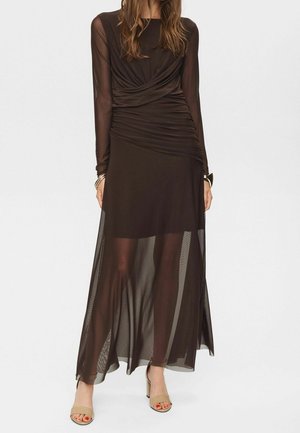 Femme portant une longue robe noire transparente avec des détails froncés et des manches longues, associée à des talons ouverts couleur nude.