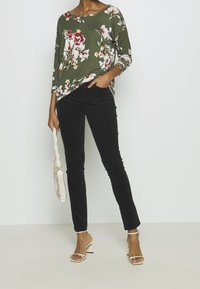 Kvinna iförd en olivgrön blommig långärmad topp, svarta skinnybyxor, vita klacksandaler, hållandes en vit vävd handväska.