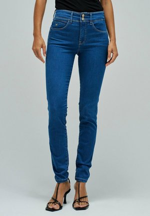 Mujer con jeans ajustados azul de talle alto con cierre de doble botón y sandalias negras de tacón alto con tiras, de pie contra un fondo gris.