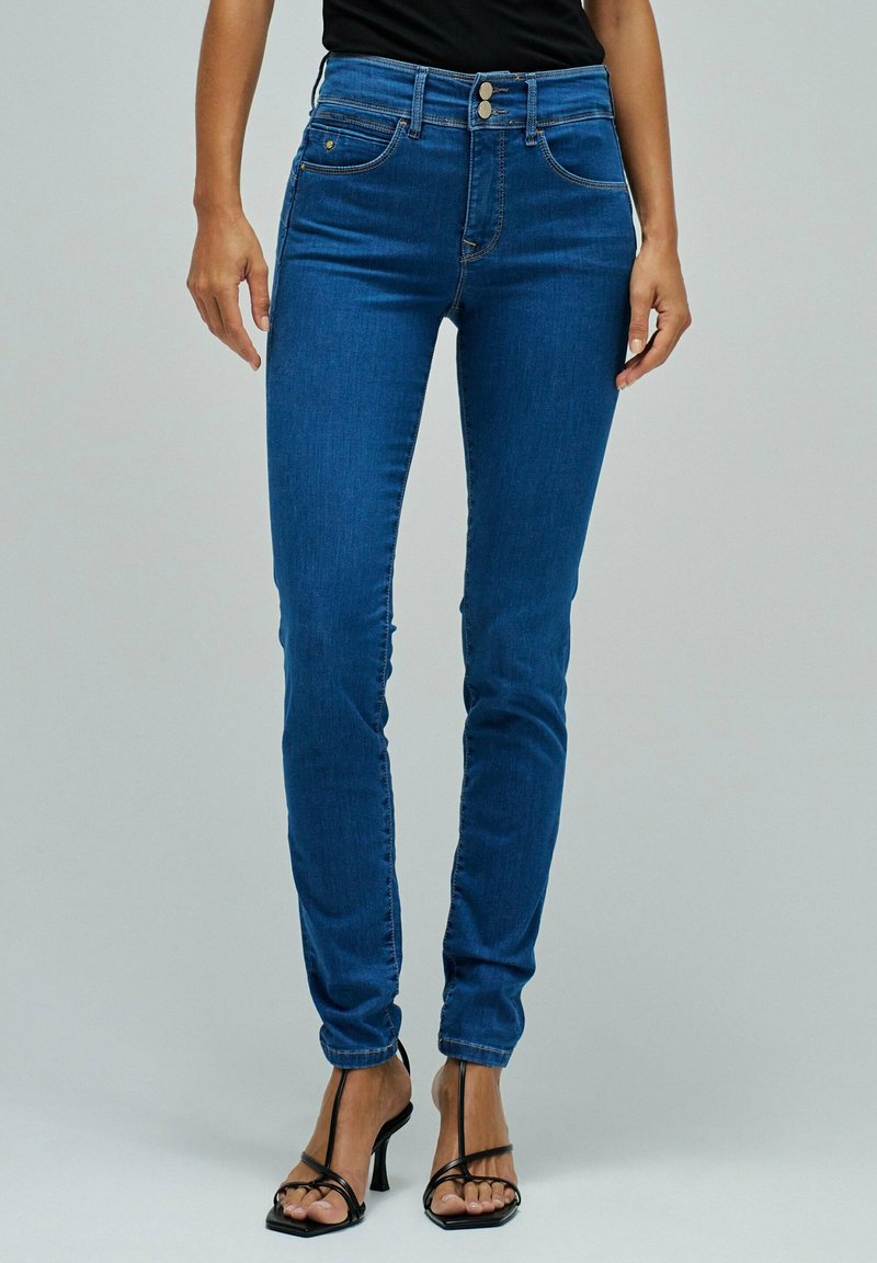 Woman wearing high-waist blue skinny jeans with double button closure and black strappy high-heeled sandals, standing against gray background.