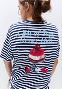 Stribet t-shirt med blå og hvide vandrette striber, med trykt dessertdesign og tekst: "En smule sukker skader aldrig" over en gelé dessert og kirsebær.