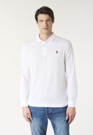 Uomo che indossa una polo bianca a maniche lunghe con un piccolo logo scuro sul petto, abbinata a jeans blu, in piedi davanti a uno sfondo chiaro e uniforme.