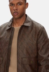 Giacca marrone a motivi con zip frontale, accenti di logo prominenti, due tasche a fessura e una finitura a superficie liscia. Maglietta beige a maglia visibile.