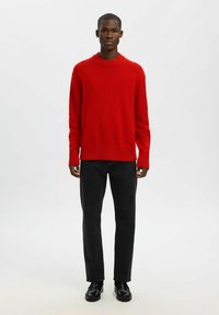 Junger Mann steht da, trägt einen leuchtend roten Pullover, schwarze Jeans und schwarze Schuhe, vor einem einfachen weißen Hintergrund.