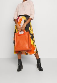 Sac cabas en cuir orange avec logo "VALENTINO" embossé, associé à une jupe colorée à motifs floraux et des bottines noires.