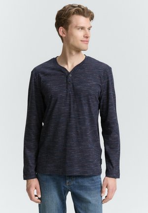 Camisa henley de manga larga en azul marino con un patrón texturizado, tapeta de tres botones y detalles de hilo en contraste, combinada con jeans azules.