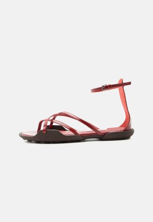 Red flat sandal with multiple thin straps, ankle buckle, and a slightly elevated brown sole against a plain light background.