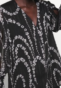 Blusa nera con motivo floreale bianco, dotata di scollatura a V, maniche trasparenti e tessuto testurizzato. Etichetta del designer visibile all'orlo.
