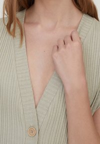 Primer plano del torso superior con una blusa beige de canalé con botones, mostrando las clavículas, una mano cerca del pecho y mechones de cabello castaño claro.