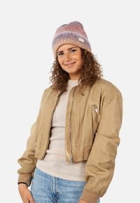 Tan bomber jacket with ribbed cuffs, beige sweater underneath, and a multi-colored knit beanie with a label. Curly hair visible.