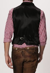 Schwarze Satin-Weste mit glatter Textur, ausgestattet mit einem Schnallen-Detail auf der Rückseite. Kombiniert mit einem rot-weiß karierten Hemd und braunen Lederhosen.