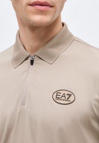 Κοντινό πλάνο ενός άνδρα που φοράει ένα μπεζ πόλο με φερμουάρ, με υφασμένο γιακά και λογότυπο EA7 Emporio Armani στο στήθος.