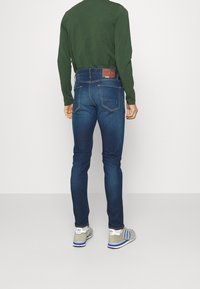 Tmavě modré džíny z denimu s přiléhavým střihem, zadními kapsami a detaily hnědého koženého záplatu u pasu; nošené s zelenou košilí s dlouhým rukávem.