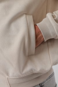 Main dans la poche d'une veste beige avec poignets et taille côtelés, montrant une texture de tissu douce et lisse.