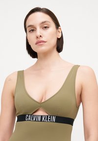 Olívazöld fürdőruha mély V-nyakkivágással és keresztezett elülső kialakítással. Fekete elasztikus szalag díszíti, amelyen a "CALVIN KLEIN" felirat látható a deréknál.