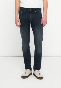 Osoba nosząca ciemnoniebieskie obcisłe dżinsy i białe trampki, stojąca na tle jednolitego białego tła.