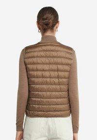 Brun pufferväst med vertikal quiltad mönster, hög krage och lätt material, bärs över en beige långärmad topp.