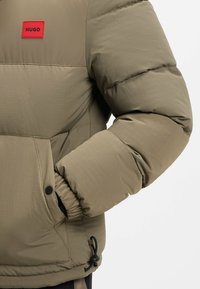 Giacca puffer in verde oliva con texture trapuntata. Presenta un'etichetta brandizzata rossa, polsini elastici e orlo regolabile.