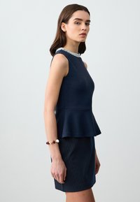 Top peplo blu navy con colletto decorato con perline, realizzato in tessuto elasticizzato. Indossato con una gonna a righe sottili. Modello inquadrato di lato, mentre tiene un bracciale.