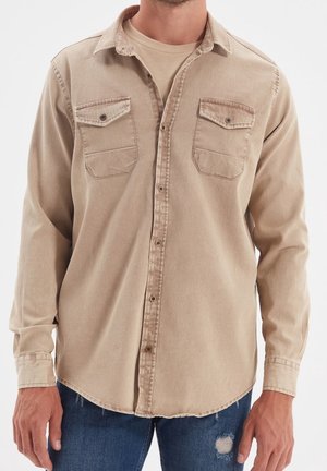 Mężczyzna ubrany w beżową koszulę na guziki z dwoma zapinanymi na guziki kieszeniami na piersi, nałożoną na beżowy t-shirt, w połączeniu z niebieskimi rozdartymi dżinsami.