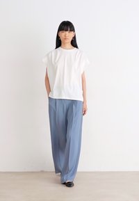 Top bianco a maniche corte abbinato a pantaloni blu larghi e scarpe nere. Tessuto liscio con vestibilità rilassata, in piedi davanti a uno sfondo uniforme.
