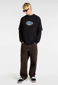 Zwart oversized T-shirt met een kleurrijk "VANS" logo, gecombineerd met losse donkerbruine jeans en klassieke zwart-witte sneakers.