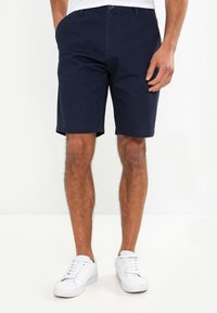 Shorts en coton bleu marine avec une coupe droite, fermeture à bouton et poches latérales. Portés avec des baskets blanches sur un fond neutre.