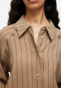 Donna che indossa una camicia beige con bottoni, a strisce verticali sottili marroni e colletto ampio, capelli sciolti e ricci.