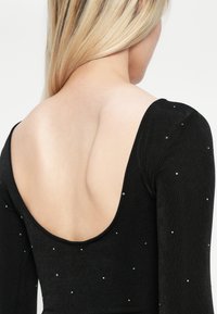 Zwarte kleding met lange mouwen en een diepe rugnek. Versierd met kleine, glanzende accenten die gelijkmatig over de stof zijn verdeeld.