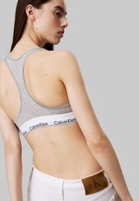 Top sportivo grigio in cotone con design a schiena scoperta e fascia bianca con logo del marchio nero, abbinato a jeans bianchi con etichetta marrone.