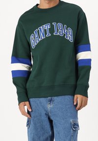 Sweatshirt vert avec "GANT 1949" imprimé en bleu, présentant des rayures bleues et blanches sur les manches. Matière en coton, design à col rond.