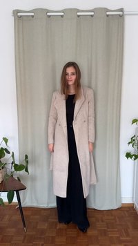 Casaco de lã bege claro com fecho duplo e colarinho de lapela, usado sobre uma roupa preta. O casaco apresenta tecido texturizado e mangas compridas.