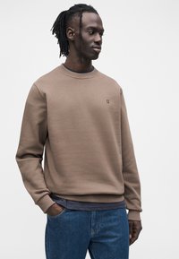 Καφέ φούτερ με ριμπ γιακά και μανσέτες, διακοσμημένο με ένα μικρό κεντημένο λογότυπο στο στήθος. Φοριέται πάνω από ένα σκούρο μπλε πουκάμισο και μπλε τζιν.