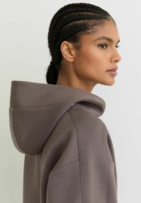 Sudadera con capucha en tono topo apagado, con tela suave, ajuste relajado y capucha prominente. Bolsillo lateral visible, textura lisa.