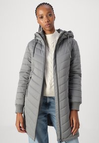 Grå quiltet boblejakke med hette, med glidelås og ribbestrikkede mansjetter. Kombinert med en kremhvitt kabelstrikkegenser og blå jeans.