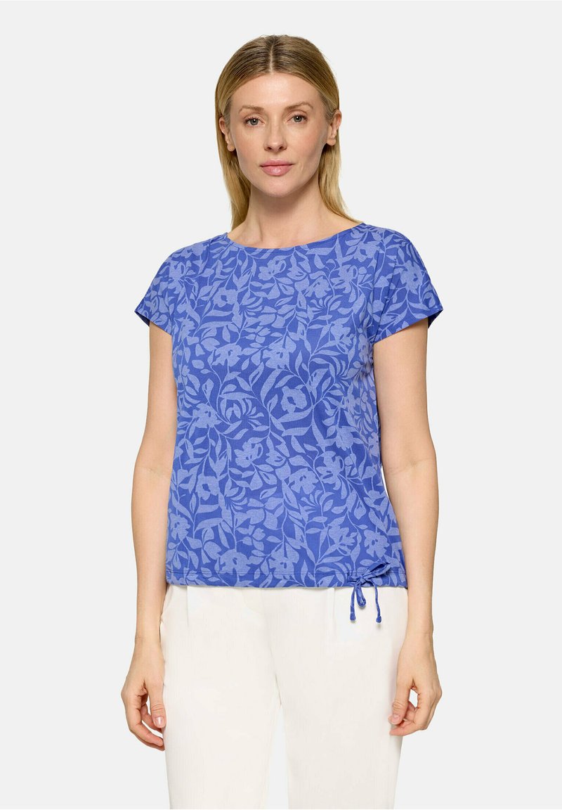 Femme portant un haut bleu à manches courtes à motif floral avec un détail noué sur le côté, assorti à un pantalon blanc sur un fond uni.