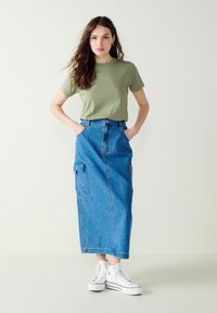 Zelené tričko s krátkym rukávom kombinované s dlhou modrou denims šatou so cargo vreckami, doplnené o biele tenisky s vysokým strihom.