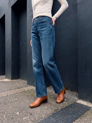 Person trägt blaue Jeans mit geradem Bein, ein beiges langärmliges Oberteil und braune Knöchelstiefel und steht draußen vor einer dunklen Wand.
